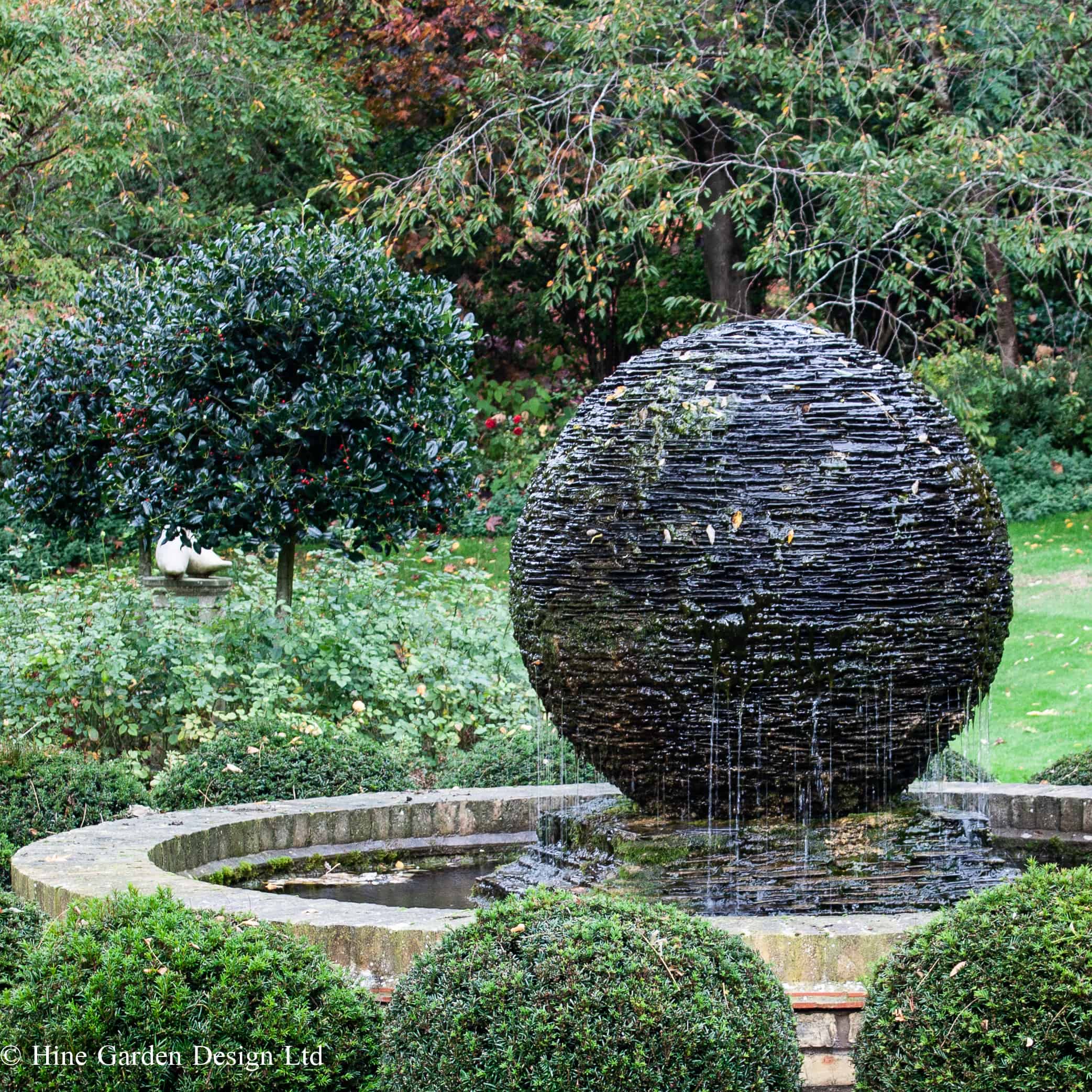 spherical dark grey stone water feature made of layered slate in a garden surrounded by topiary in a pool